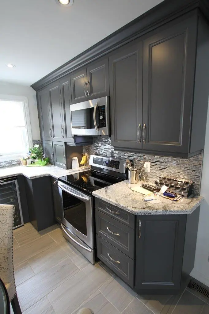 Modern kitchen with dark gray cabinets, stainless steel appliances including a microwave and electric stove, light granite countertops, mosaic tile backsplash, and light gray floor tiles.