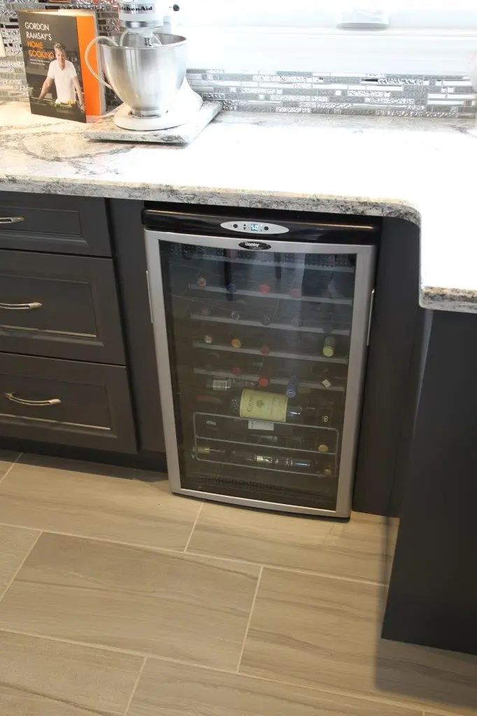 Built-in wine cooler with a digital temperature display below a light granite countertop, surrounded by dark grey kitchen cabinets and drawers, with a stand mixer and a cookbook on the counter above it.