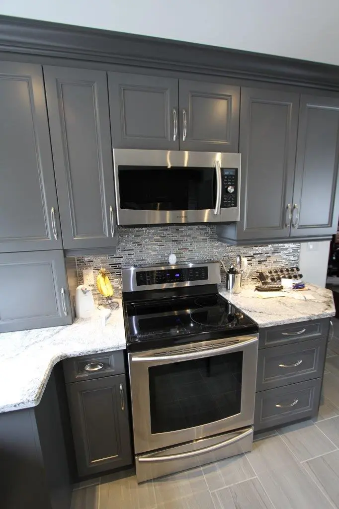 Modern kitchen setup with gray cabinets, a stainless steel microwave above a stainless steel electric stove with a black glass cooktop, a marble countertop with kitchen accessories including a banana holder, a wine rack, and a small appliance, and a mosaic tile backsplash.