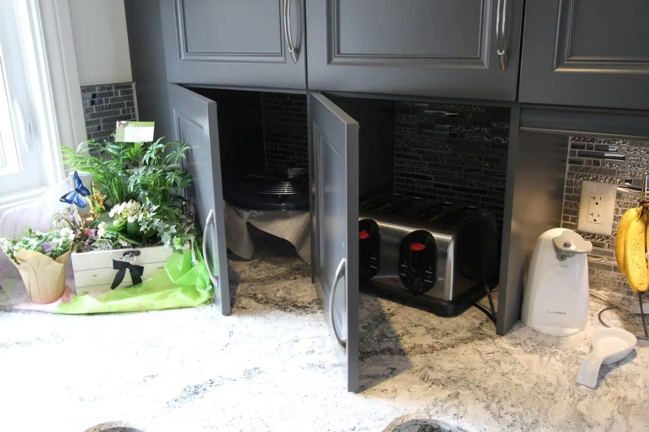 Kitchen counter with two open gray cabinets revealing a toaster and a covered dish, next to a white automatic soap dispenser, hanging bananas, a white spoon rest, and a white wooden crate filled with various green plants and flowers beside a window.