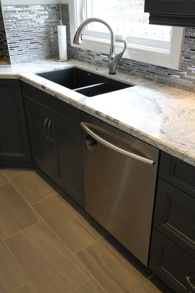 Modern kitchen countertop with a black undermount sink, stainless steel faucet, stainless steel dishwasher, dark cabinetry, gray marble-like surface, and silver mosaic tile backsplash beneath a window.