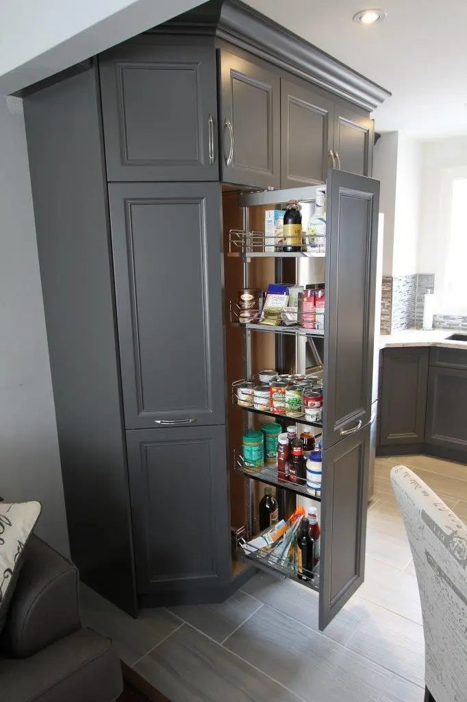 Tall gray kitchen pantry cabinet with one door open showing pull-out metal shelves stocked with various canned goods, bottles, and containers, next to a bright kitchen corner with countertop and dark lower cabinets.