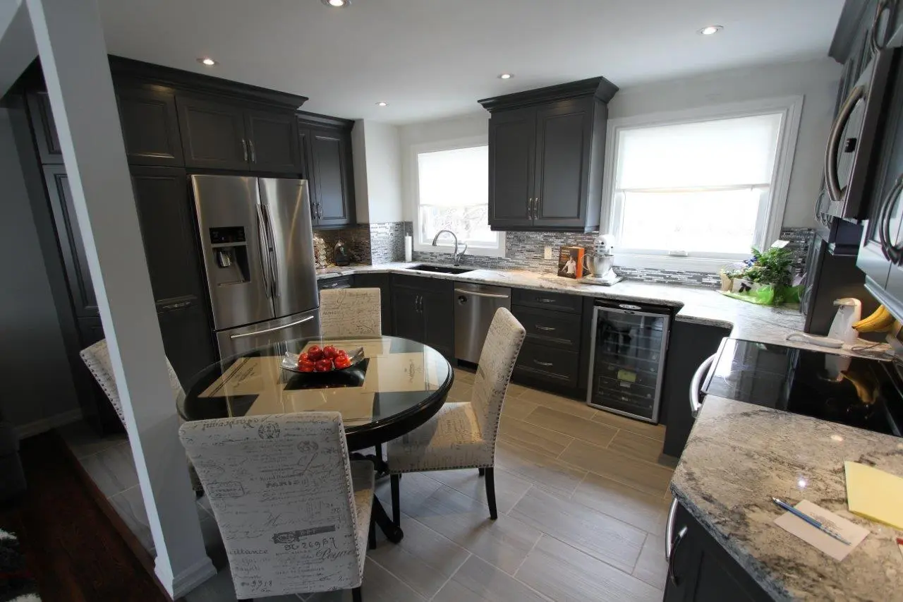 Modern kitchen with dark gray cabinetry, stainless steel appliances including a refrigerator, dishwasher, and wine cooler, a round glass dining table with upholstered chairs featuring script patterns, and light gray tile flooring.