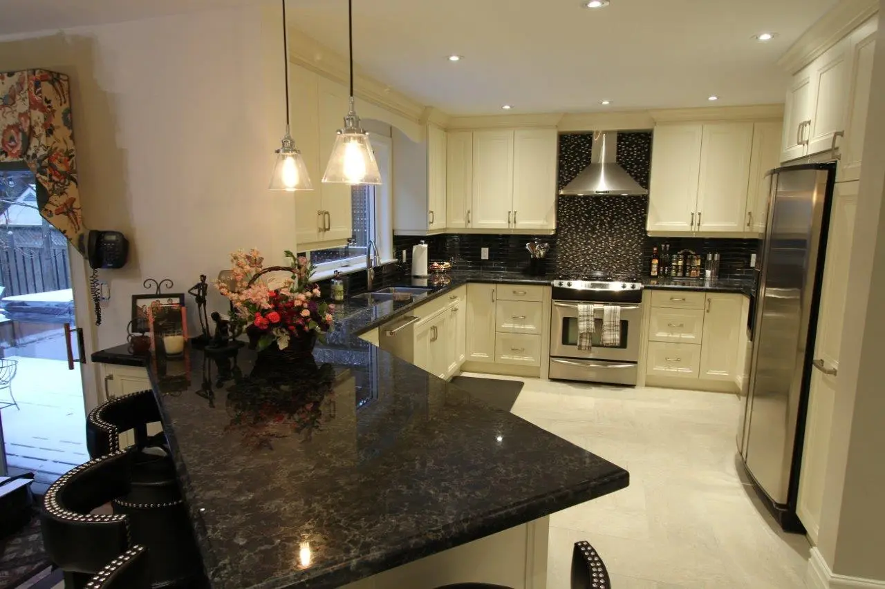 Modern kitchen with cream-colored cabinets, black granite countertops, stainless steel appliances including a stove and refrigerator, a floral basket centerpiece on the counter, and pendant lights hanging from the ceiling.