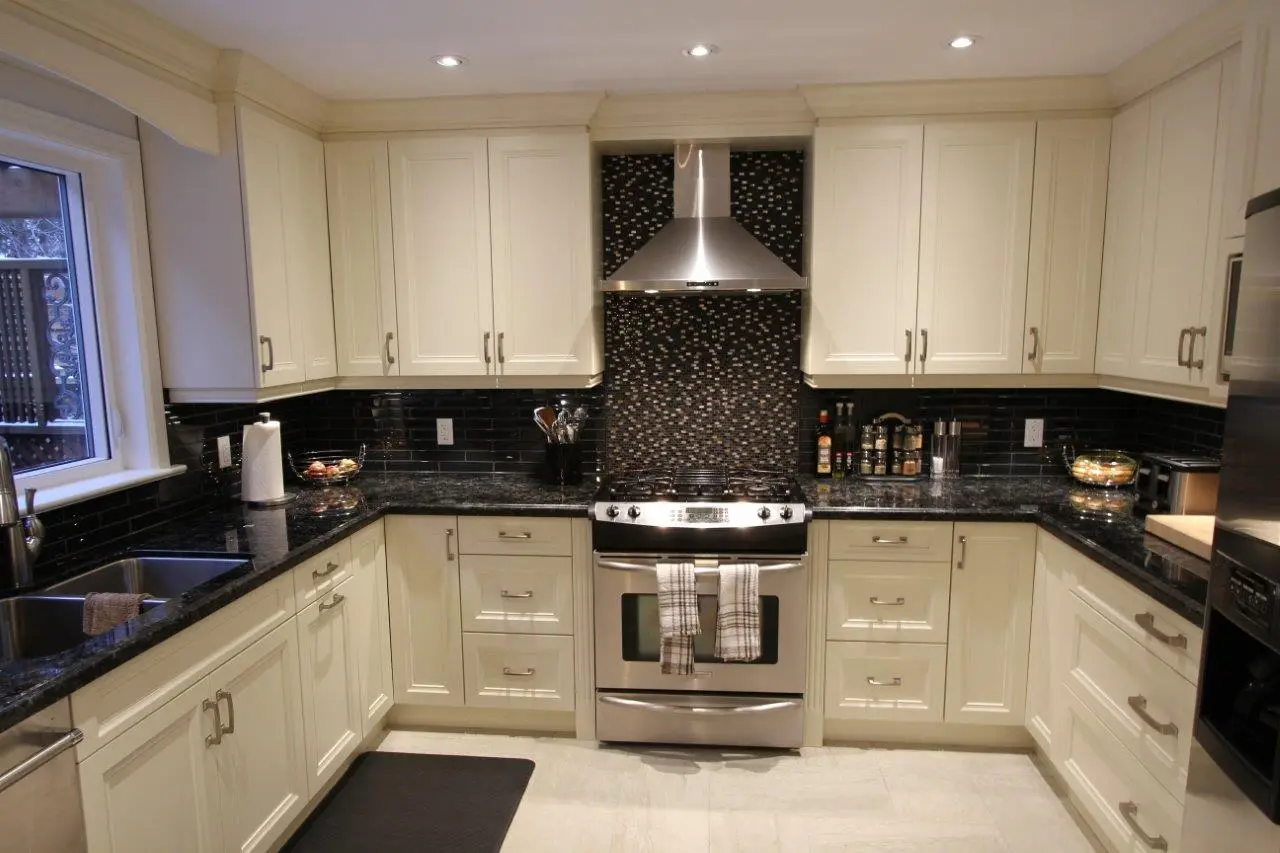 Modern kitchen with cream-colored cabinets, black granite countertops, a stainless steel stove with two kitchen towels hanging on the oven door, a matching stainless steel range hood, black tiled backsplash, and a double sink under a window on the left.