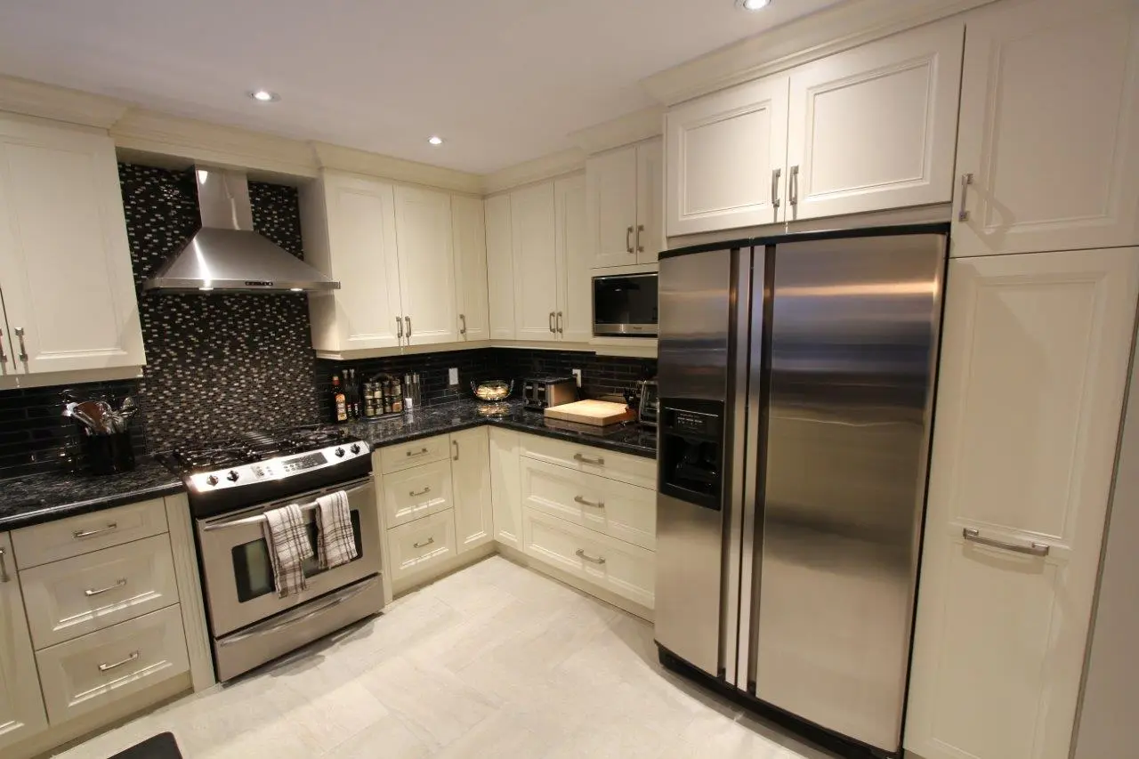 Modern kitchen with cream cabinets, black granite countertops, a stainless steel side-by-side refrigerator, a stainless steel gas stove with a hood vent, mosaic backsplash behind the stove, and small kitchen appliances on the counters.