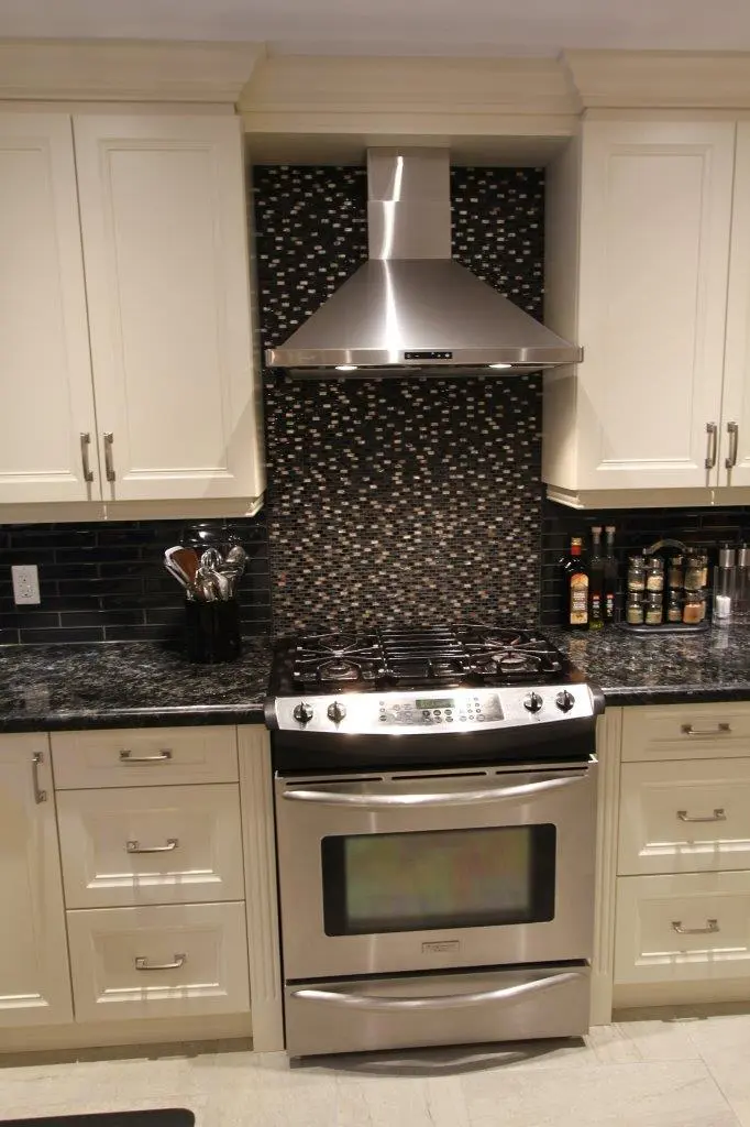 Modern kitchen with a stainless steel gas stove and oven below a stainless steel range hood, surrounded by white cabinets and a black and gray mosaic tile backsplash, with kitchen utensils and spices on the dark granite countertop.