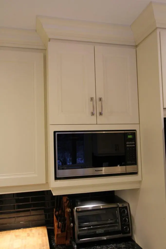 Kitchen corner with white cabinets, a built-in Panasonic microwave showing 6:00 on the display, a black toaster oven on the countertop, a wooden knife block with knives, and a black tiled backsplash.