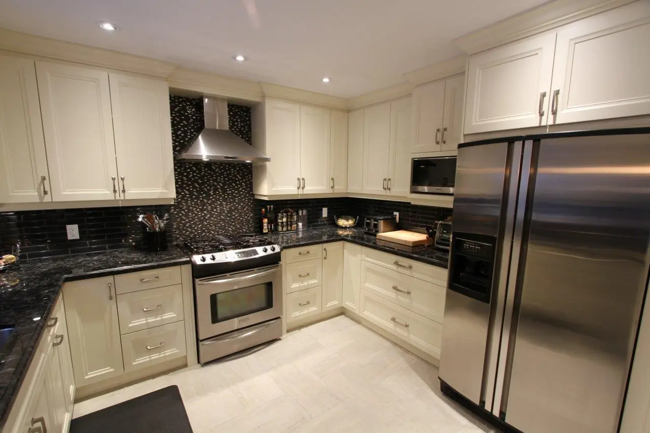 Modern kitchen with cream-colored cabinets, black granite countertops, stainless steel appliances including a double-door refrigerator and a stove with an overhead vent hood, black tile backsplash with a mosaic accent behind the stove, and light-colored tiled floor.