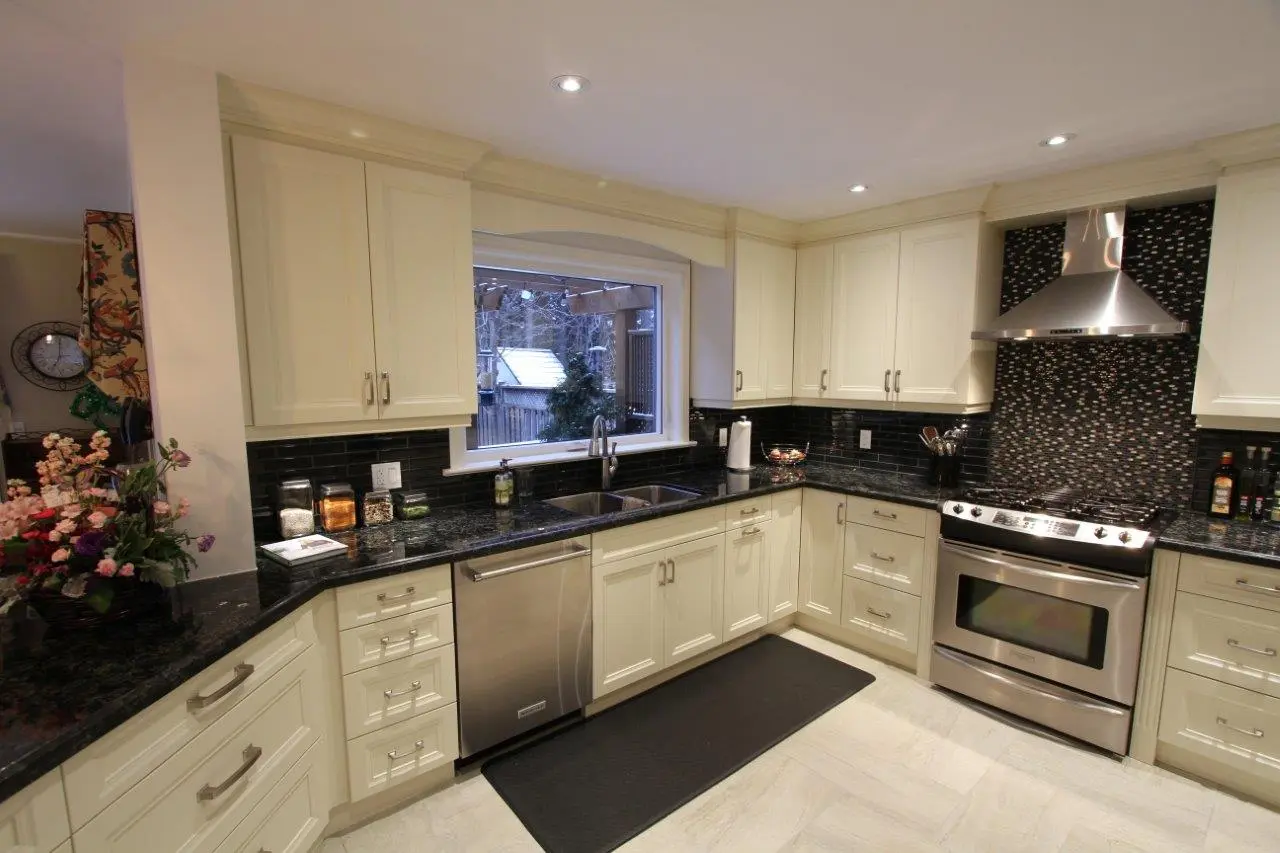 Modern kitchen with cream cabinets, black granite countertops, black tile backsplash, stainless steel appliances including a dishwasher and stove with a hood, a large window above the sink overlooking a backyard, and a black floor mat in front of the sink and stove.
