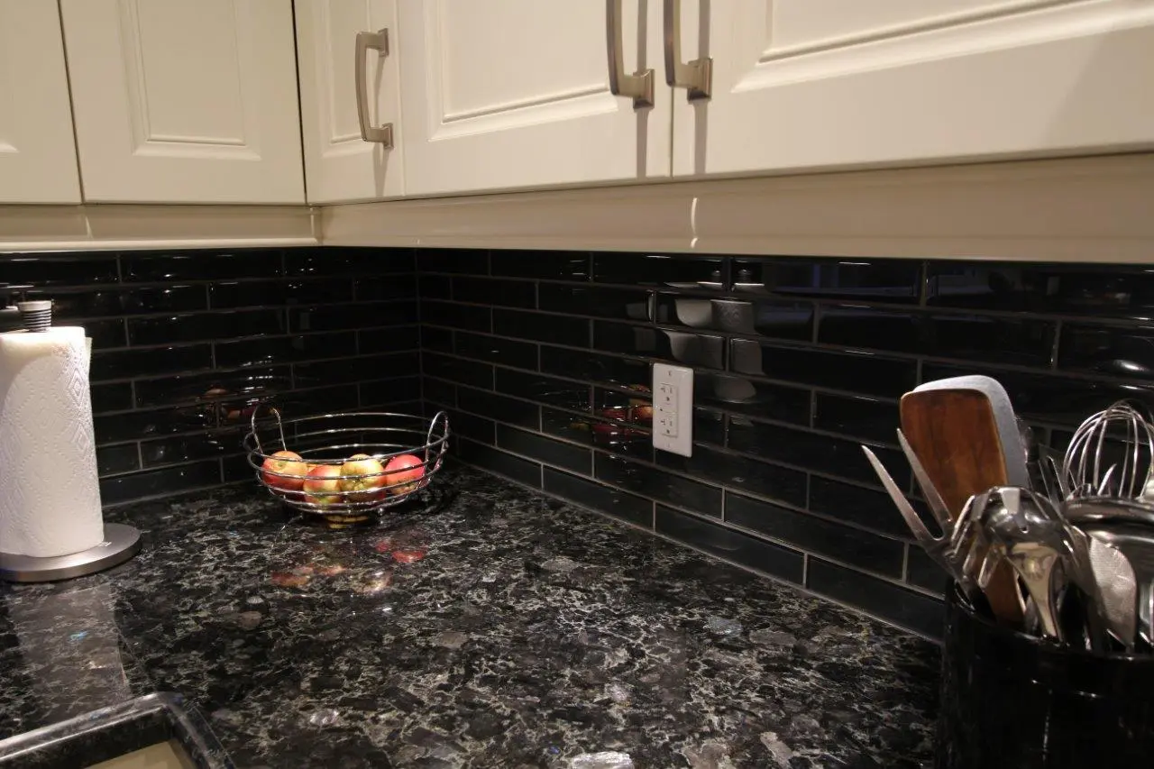 Kitchen countertop with black marble surface, black subway tile backsplash, cream-colored cabinets, a stainless steel paper towel holder with white paper towels on the left, a wire fruit basket with apples in the center, and a black utensil holder containing wooden and metal kitchen tools on the right.