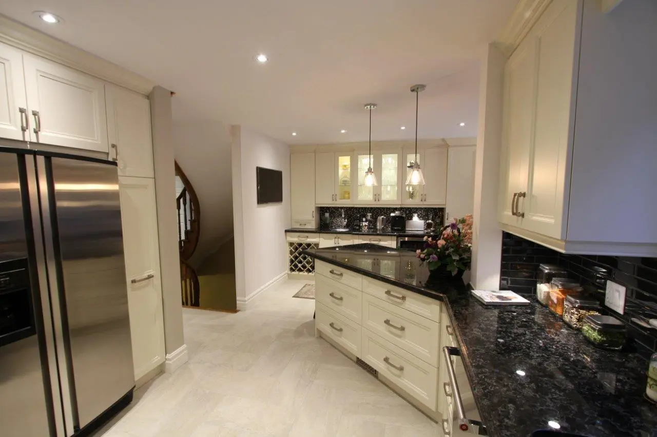 Modern kitchen with cream-colored cabinets, black granite countertops, stainless steel refrigerator, and pendant lights hanging over a corner island, with stairway visible in the background.