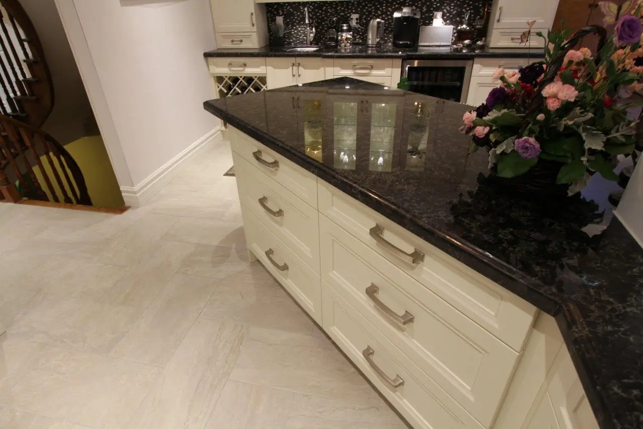 Modern kitchen interior with cream-colored cabinets, dark granite countertop island, tiled floor, and a basket of fresh flowers on the countertop near a staircase.