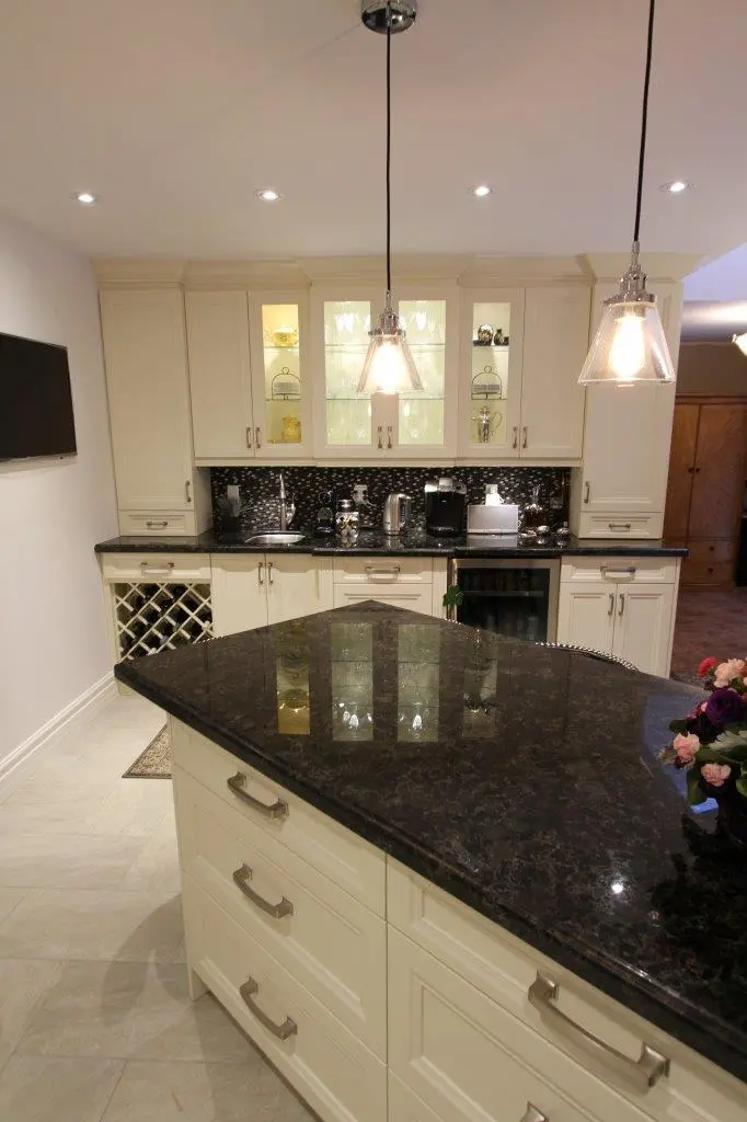 Modern kitchen featuring cream-colored cabinets with glass display sections, dark granite countertops, a central island with drawers, and pendant lights hanging from the ceiling, with various kitchen appliances on the back counter and a small vase of flowers on the island.
