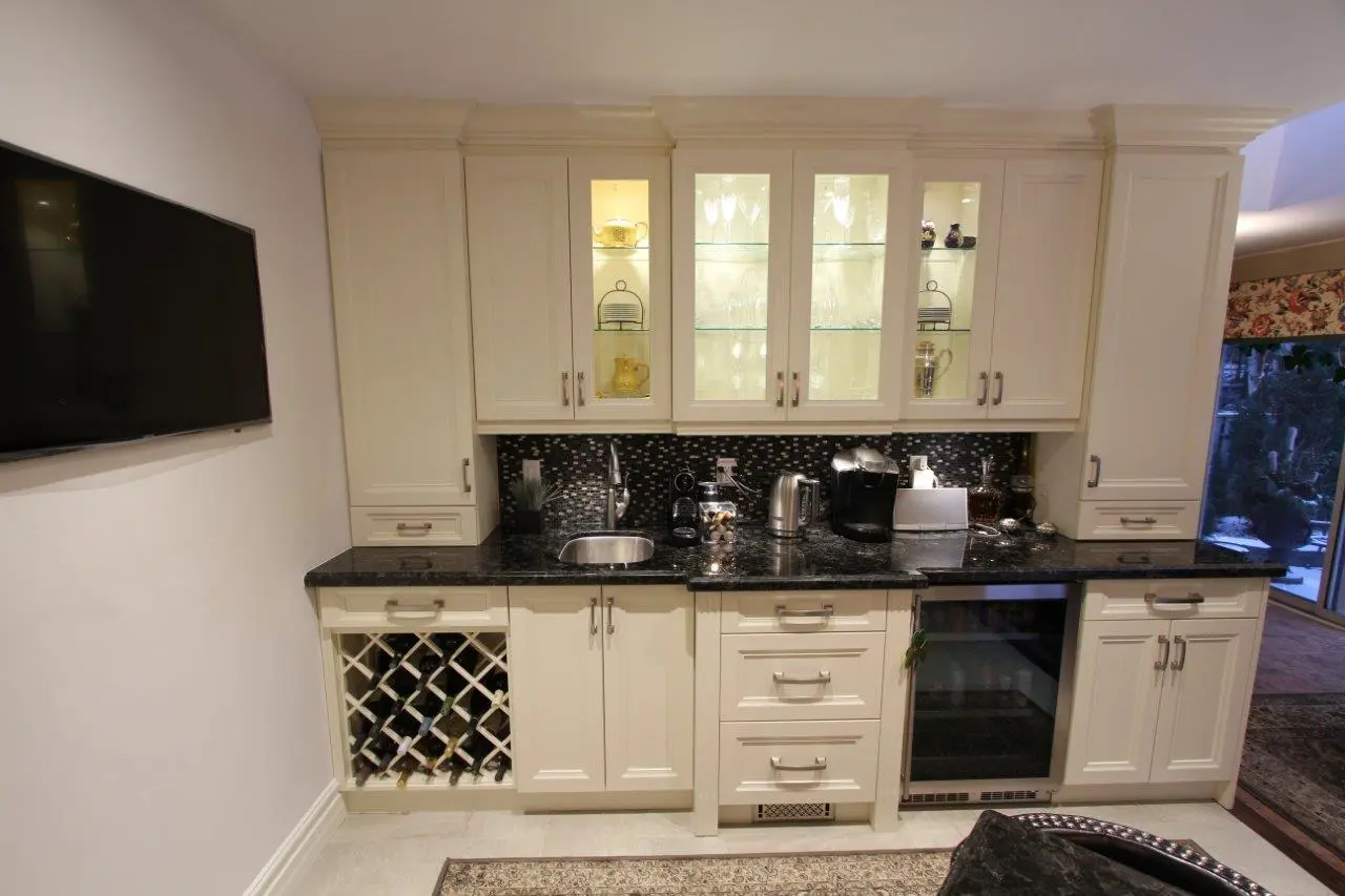 White kitchen cabinetry with black granite countertop featuring a built-in wine rack on the left, a small sink, a coffee maker, kettle, and a glass-front beverage cooler on the right, under lighted display cabinets, beside a mounted flat-screen TV on a white wall with a patterned rug on the floor.