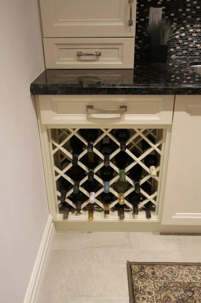 Built-in cream-colored wooden wine rack with several bottles horizontally stored, set below a black marble countertop in a kitchen with a tiled backsplash and a small plant in the corner.