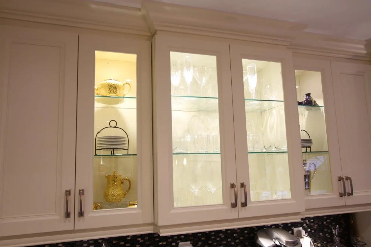 White kitchen cabinets with glass doors displaying glassware, gold-patterned teapot and pitcher, stacked plates on metal stands, and a few decorative items.