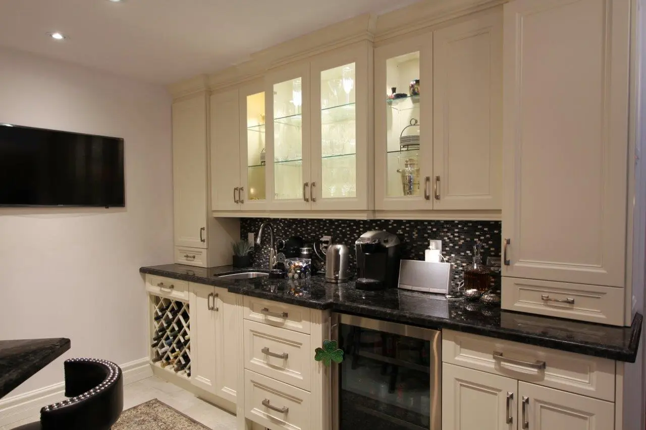 A modern kitchen corner with cream-colored cabinets, black granite countertops, a built-in wine rack filled with bottles, a small sink, coffee machine, electric kettle, and glass cabinets displaying glassware, with a flat-screen TV mounted on the adjacent wall.