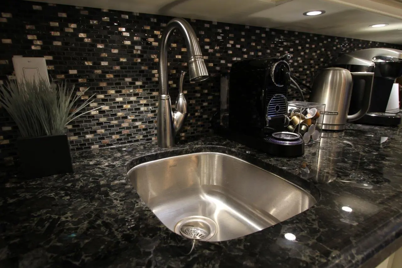 Modern kitchen countertop with a stainless steel sink, a curved faucet, a black coffee machine with coffee pods, a stainless steel electric kettle, and a decorative plant against a black and brown mosaic tile backsplash.