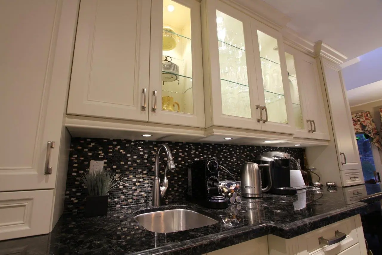 Modern kitchen countertop with black marble surface, stainless steel sink, chrome faucet, mosaic tile backsplash, white upper cabinets with glass doors displaying glassware, and kitchen appliances including two coffee makers and an electric kettle.