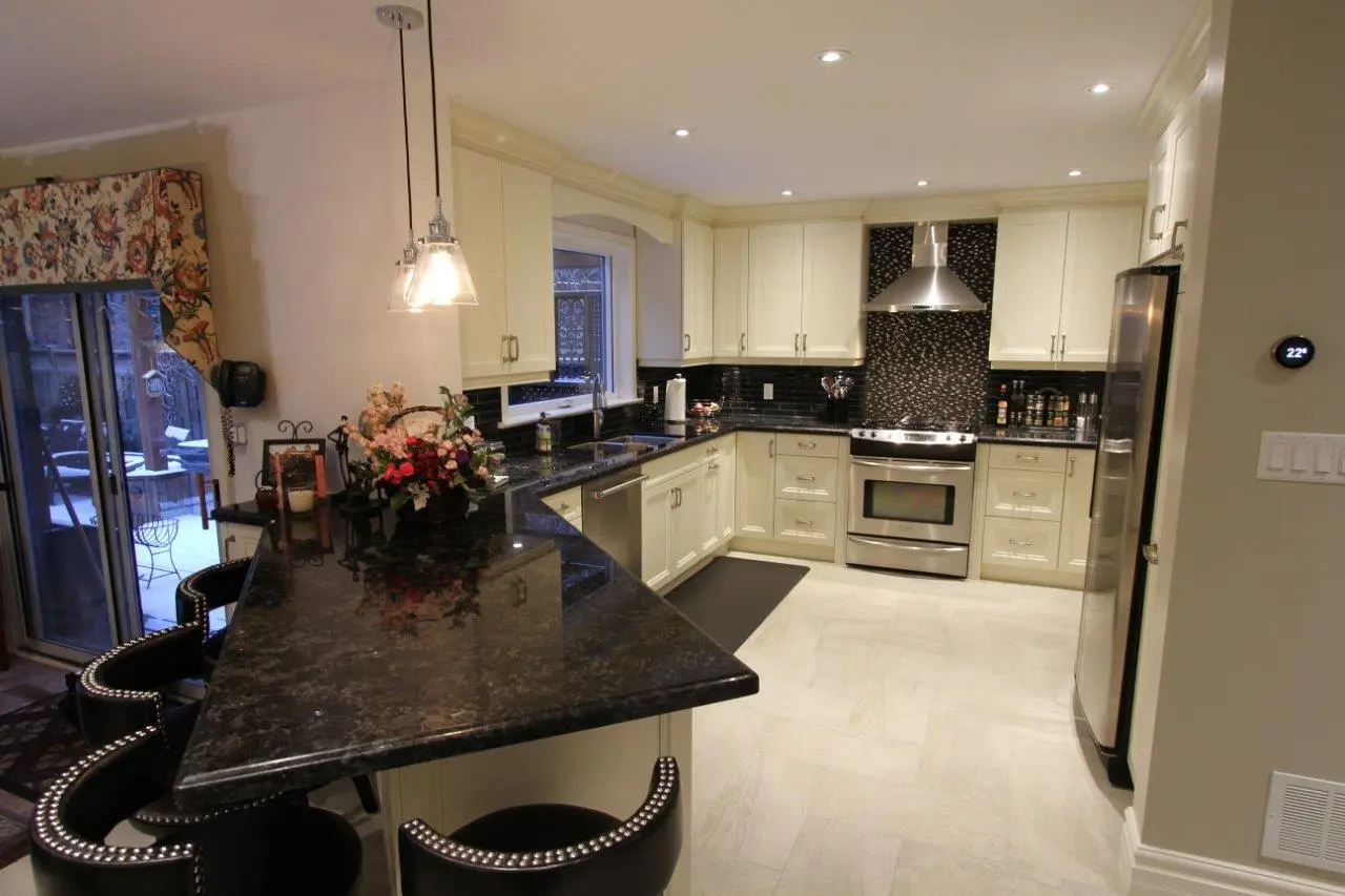 Modern kitchen with black granite countertops, cream-colored cabinets, stainless steel appliances including a stove and refrigerator, black marble backsplash behind the stove, black cushioned bar stools, and pendant lighting above the counter.