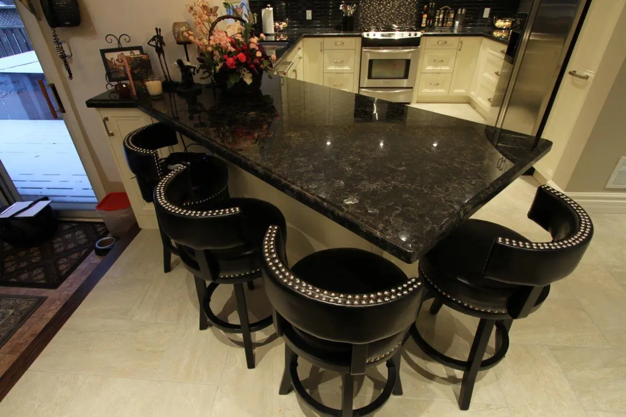 Kitchen with a black marble countertop island surrounded by five black cushioned bar stools with silver studs, cream-colored cabinets, stainless steel appliances, and a flower arrangement on the countertop.