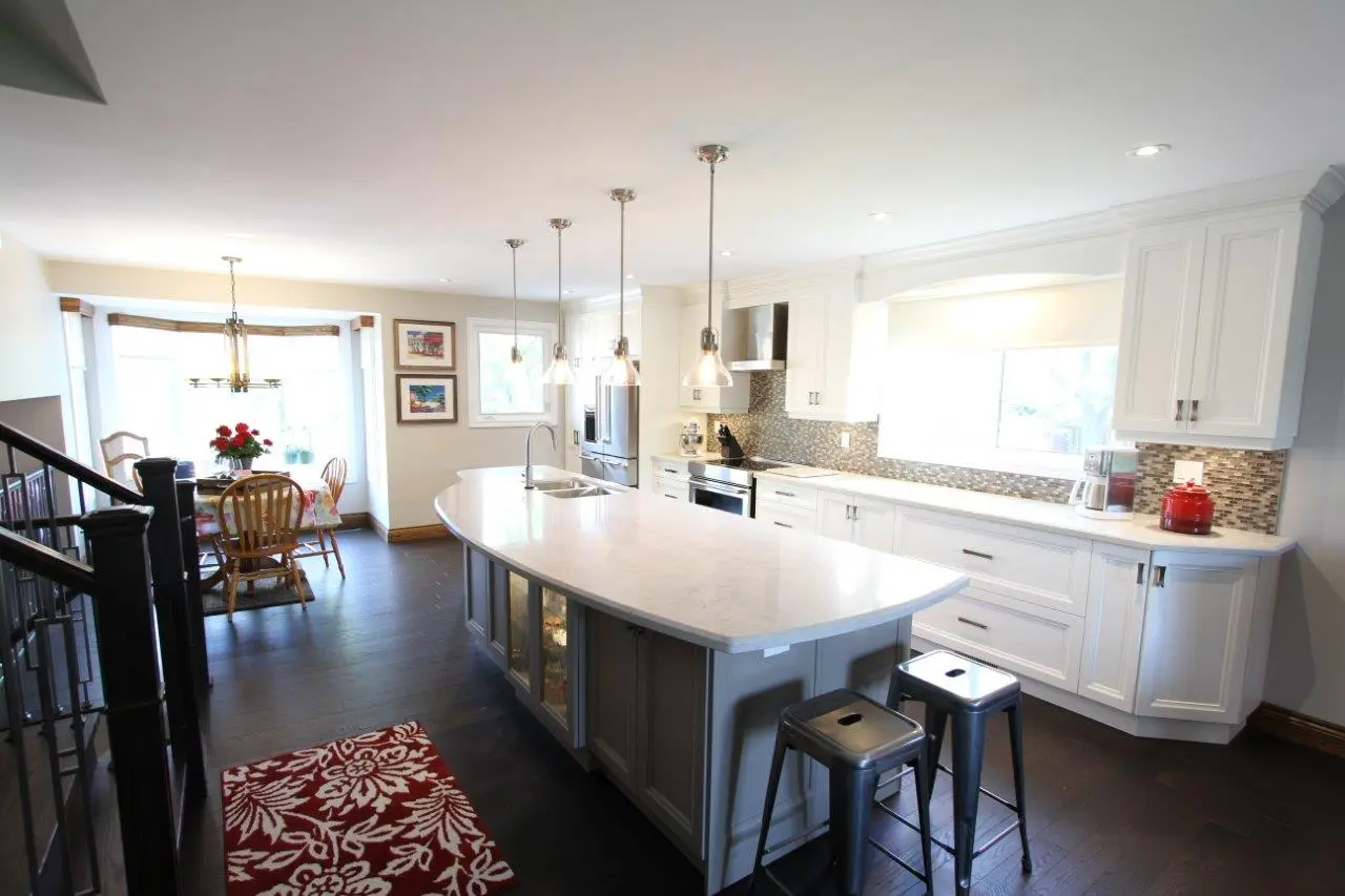 Modern kitchen with a large white marble island, two metal bar stools, white cabinets, a long countertop with mosaic backsplash, and a dining area with wooden chairs and a table near large windows.