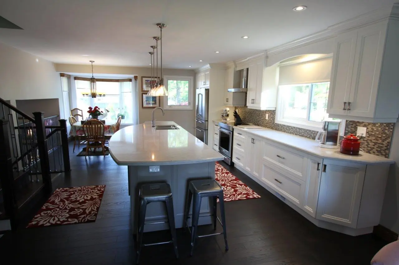 Bright modern kitchen with white cabinets, a large central island with two metal stools, dark hardwood floors, and a dining area with wooden chairs near a bay window.