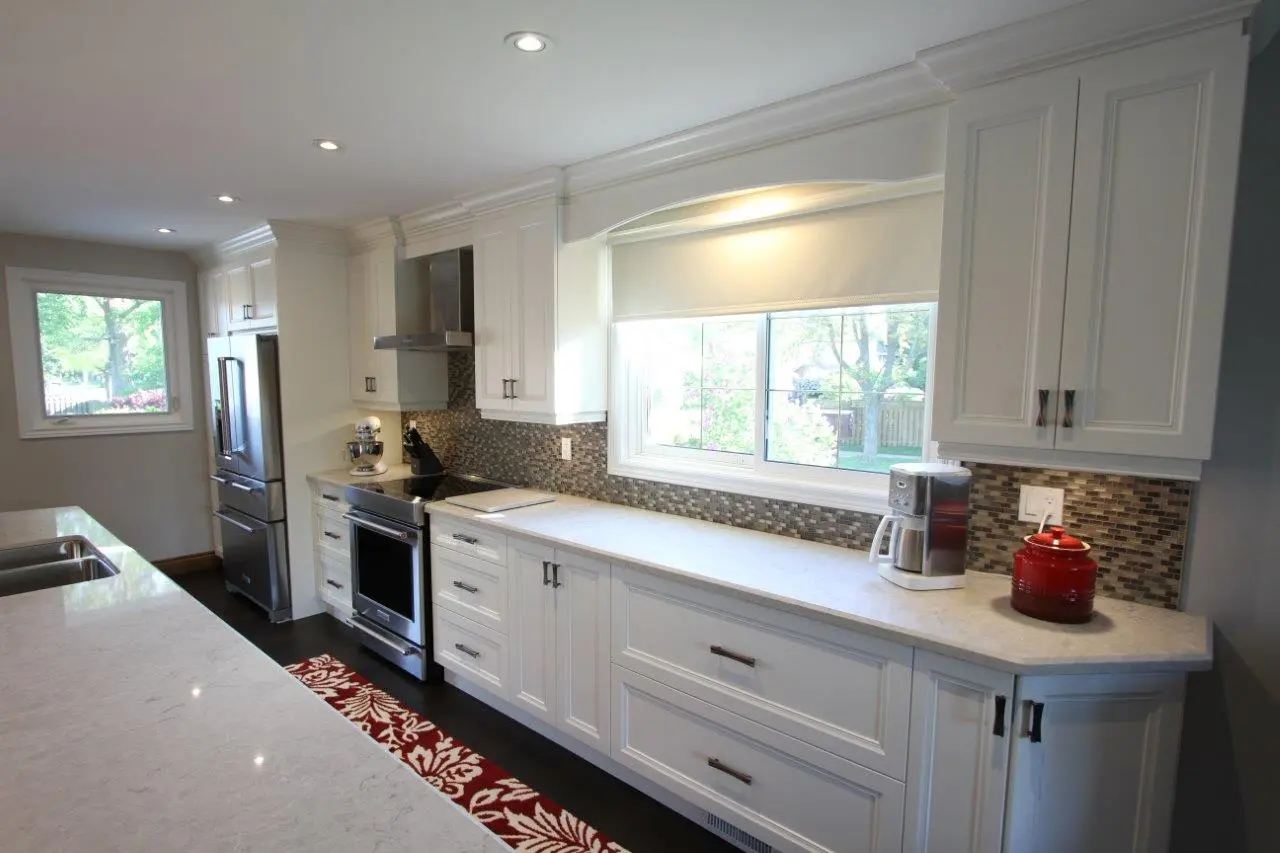 Modern kitchen with white cabinetry, stainless steel appliances, mosaic tile backsplash, large window with white shade, a coffee maker and red container on the countertop, and a red and white patterned runner rug on the dark floor.