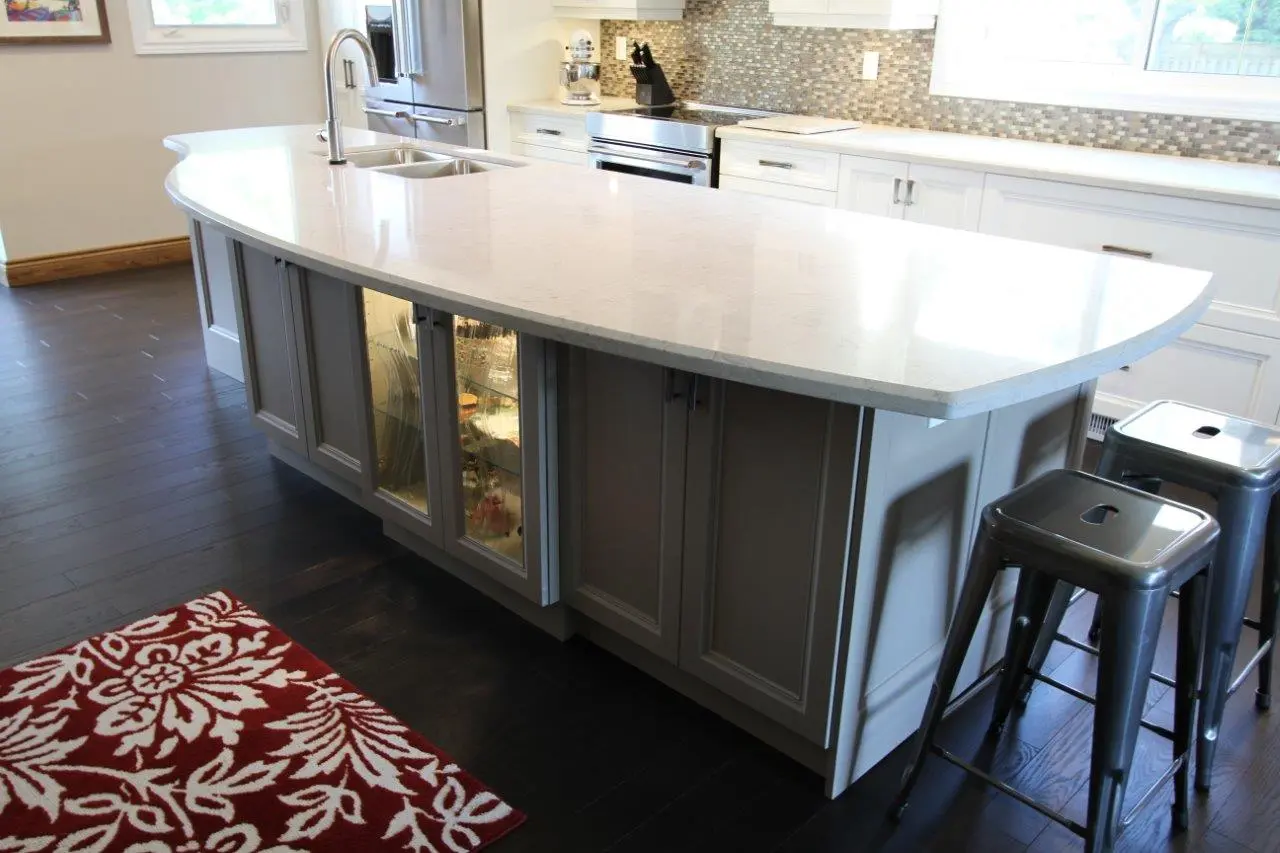 Modern kitchen island with a smooth white countertop, built-in sink with a curved faucet, gray cabinets including glass-fronted display cabinets, two metallic bar stools, dark wooden floor, and red and white floral patterned rug nearby.