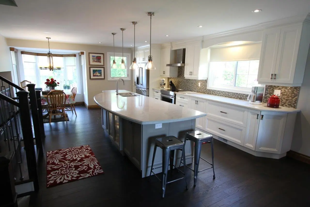Bright modern kitchen with white cabinets, a large island with a curved marble countertop, two metal bar stools, pendant lights hanging above the island, and a dining area with wooden chairs near a bay window.