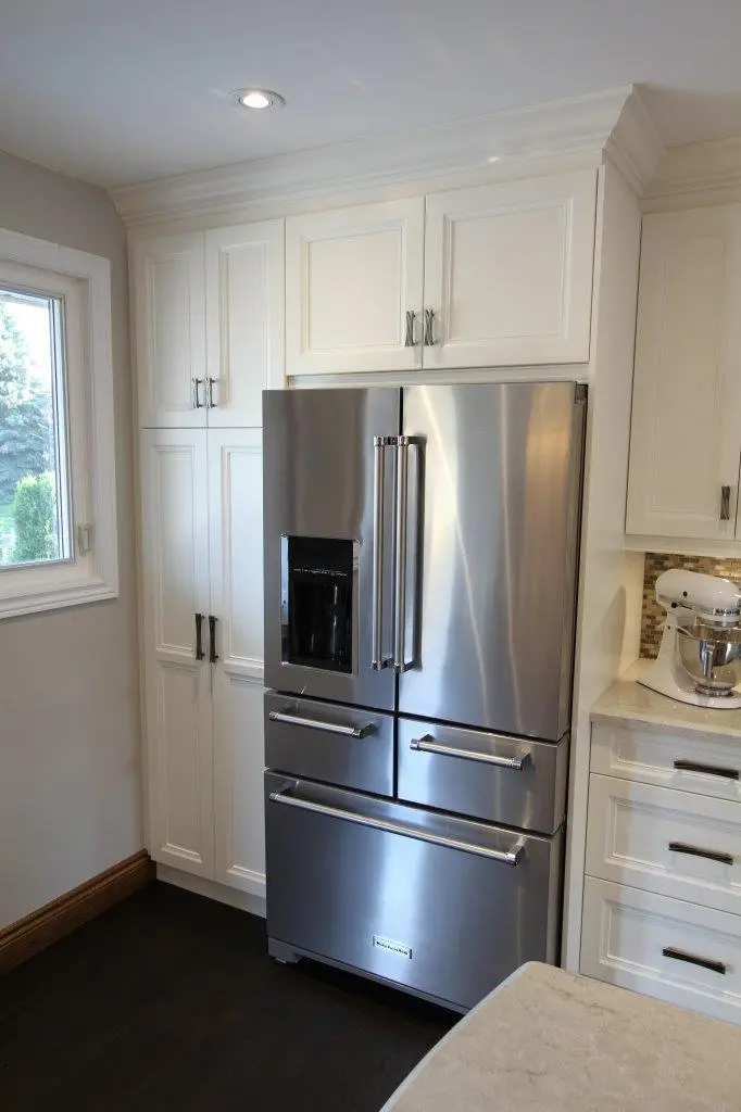 Modern kitchen corner featuring a stainless steel French door refrigerator with water dispenser, surrounded by white cabinetry and a window on the left.
