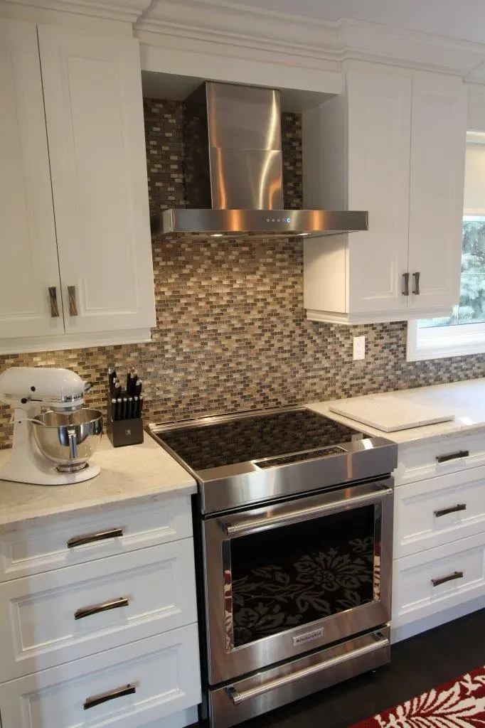 Modern kitchen corner featuring a stainless steel electric stove with a black glass cooktop, a stainless steel range hood, white cabinets, a countertop with a white mixer, a knife block, and a mosaic tile backsplash in shades of brown and beige.