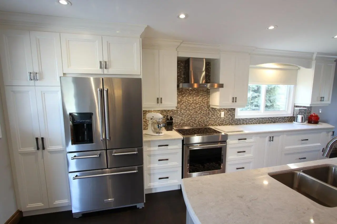Modern kitchen with white cabinetry, stainless steel French door refrigerator, built-in oven with a glass cooktop, and mosaic tile backsplash, featuring a marble countertop island with a double sink and a window above the counter letting in natural light.