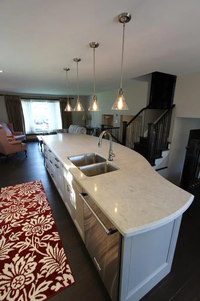 Modern kitchen with a long white marble island featuring a built-in stainless steel double sink, a dishwasher, four hanging pendant lights above, dark hardwood floor, and a red and white floral patterned rug on the left side.