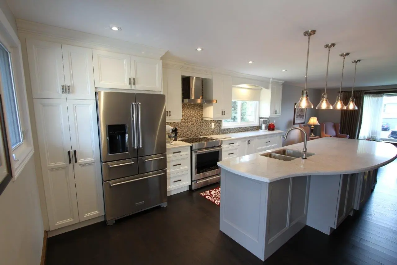 Modern kitchen with white cabinetry, a large stainless steel refrigerator, a built-in oven and stove, a mosaic tile backsplash, and a spacious white marble island with a sink and hanging pendant lights above, dark hardwood floors, and seating area visible in the background near large windows.