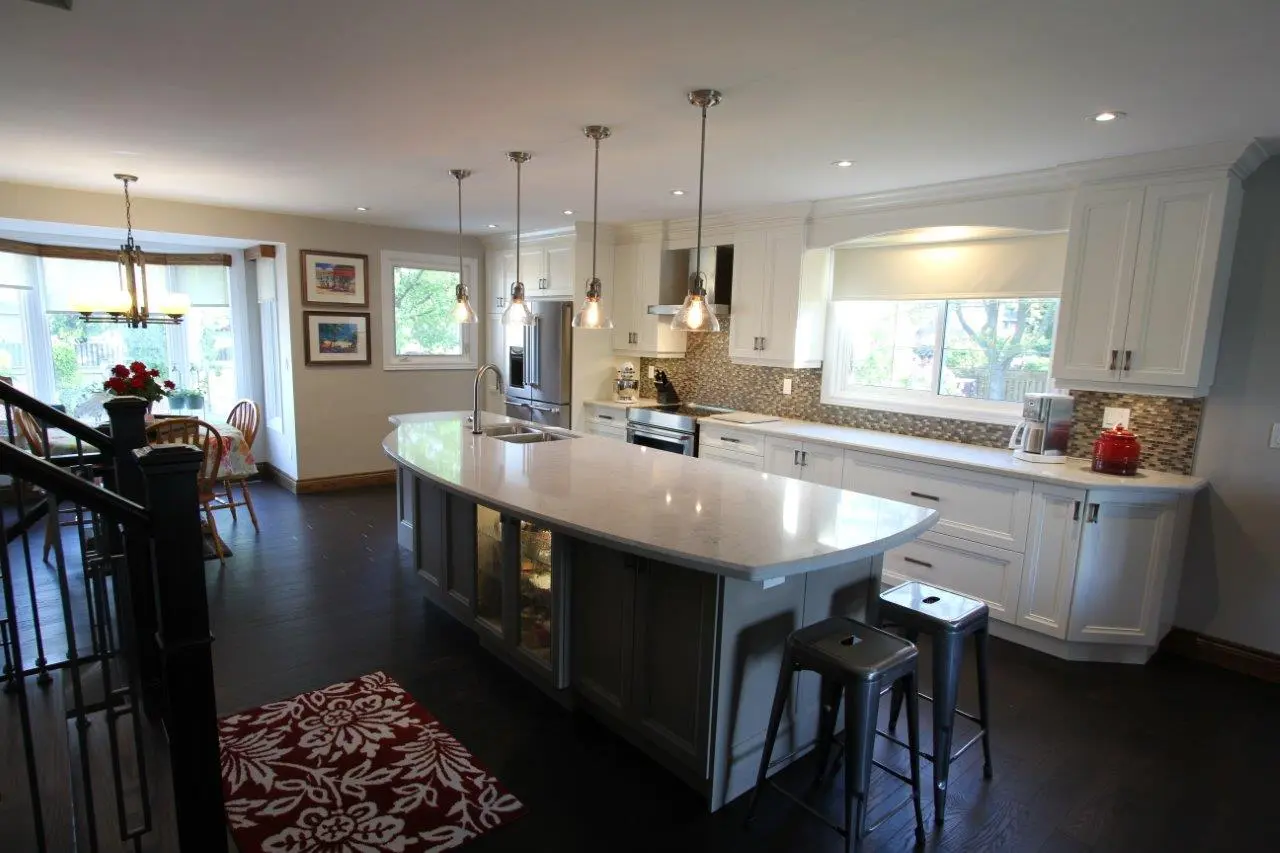 Modern kitchen with white cabinets, a large curved island with a sink and four pendant lights, two metal bar stools, dark wooden floor, dining area with wooden chairs and a table near windows, and decorative elements including red flowers and a red kettle.