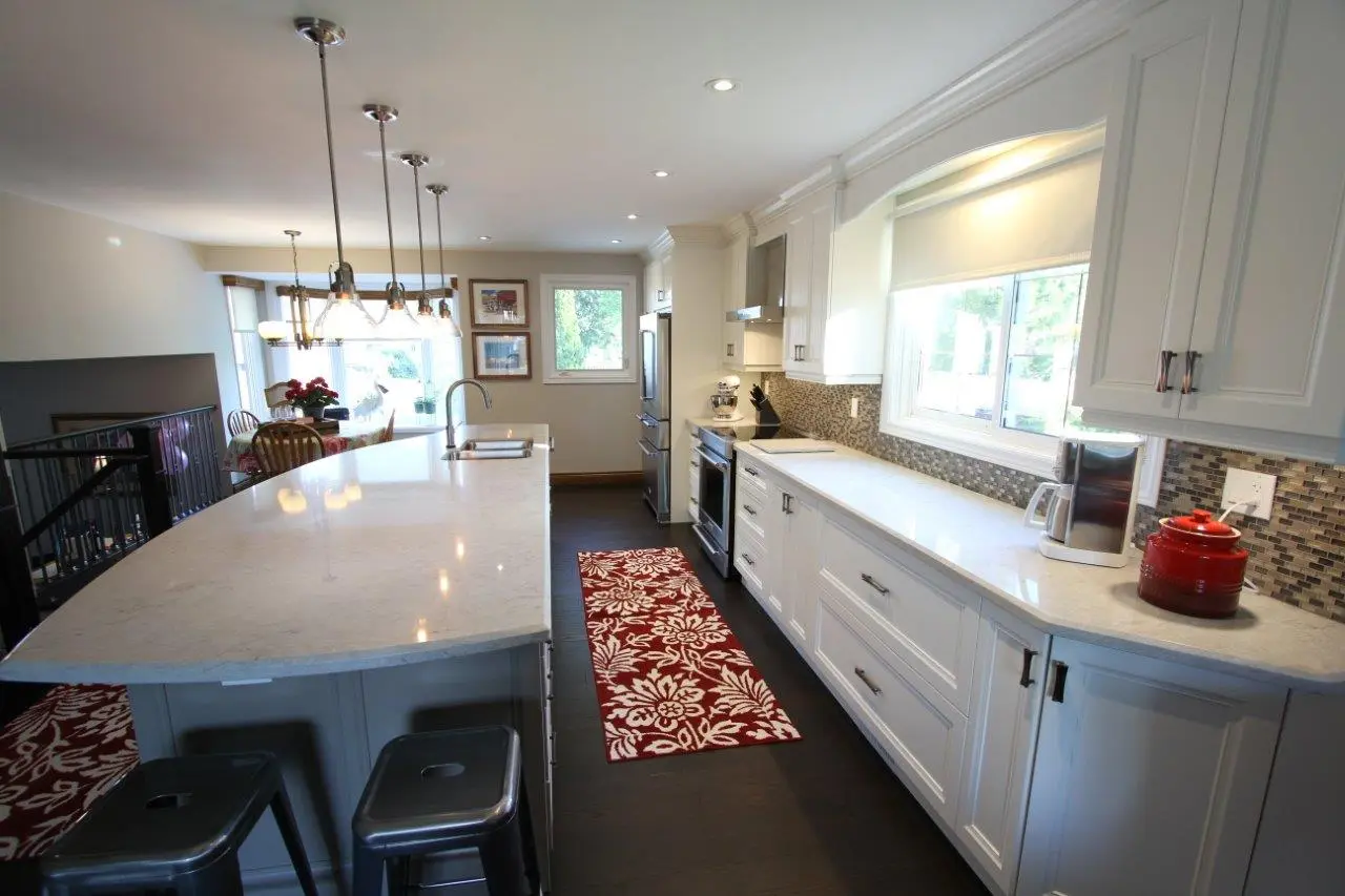 Modern kitchen featuring white cabinets, a long marble island with hanging pendant lights, stainless steel appliances, a red patterned runner rug, and a window with a roller shade letting in natural light.