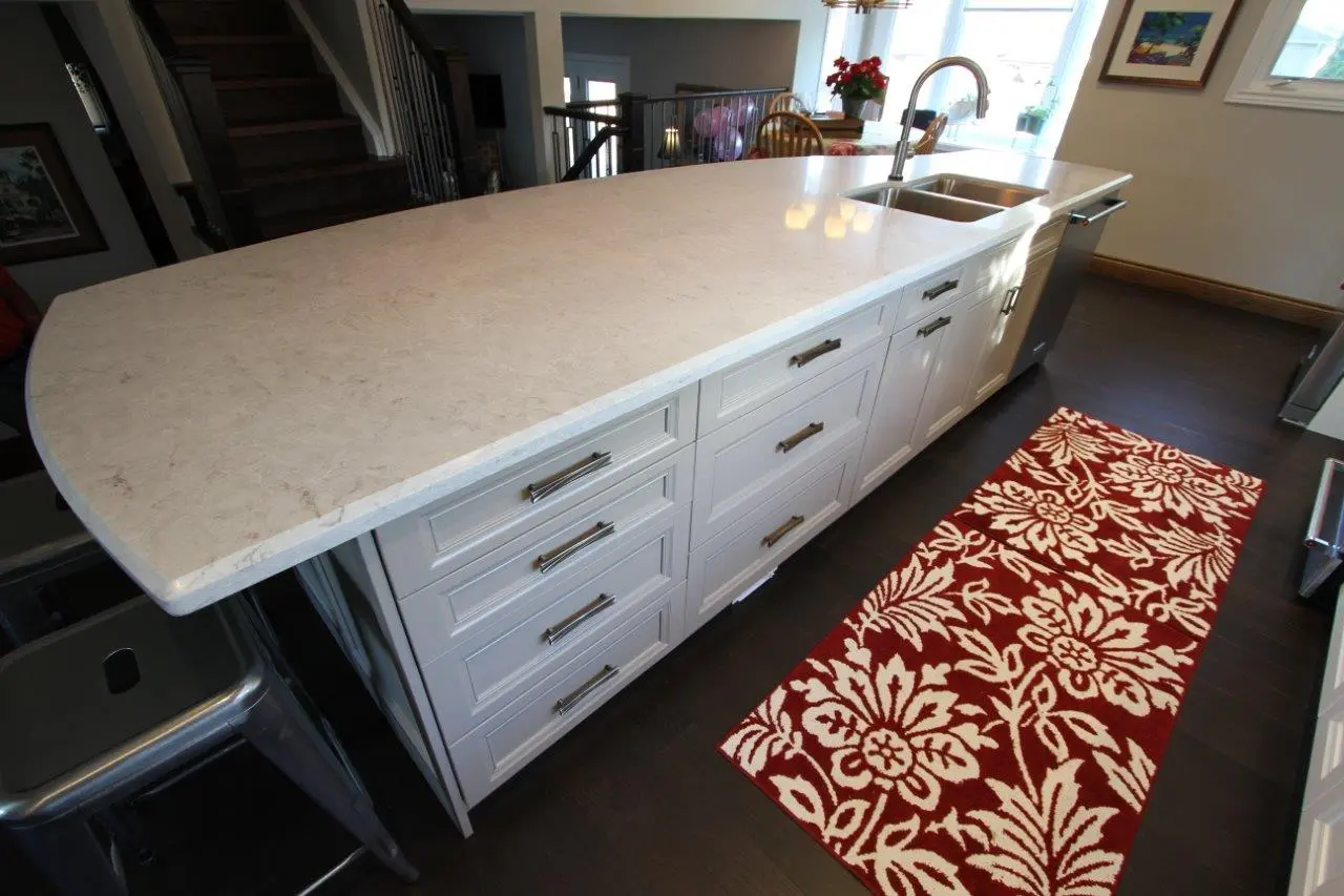 Modern kitchen island with white marble countertop, white cabinets with metal handles, built-in double sink, and a red and white floral patterned rug on dark wooden floor.