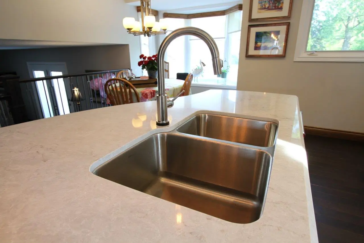 Close-up of a modern kitchen countertop with a double stainless steel sink and a sleek, curved faucet, with a dining area visible in the background featuring wooden chairs, a table with a floral tablecloth, and windows letting in natural light.