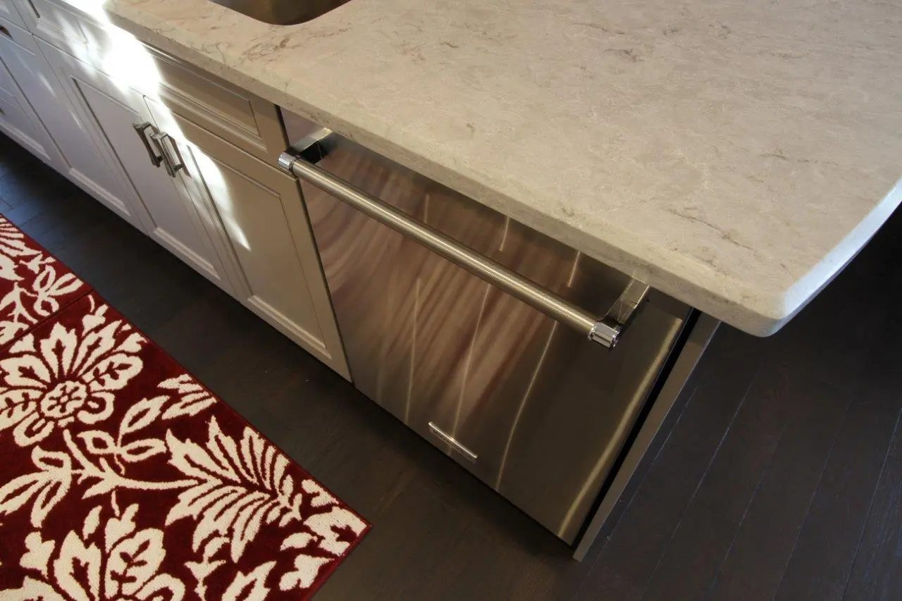Stainless steel dishwasher integrated into white kitchen cabinetry with a beige marble countertop above and a red and white patterned rug on the dark wood floor.