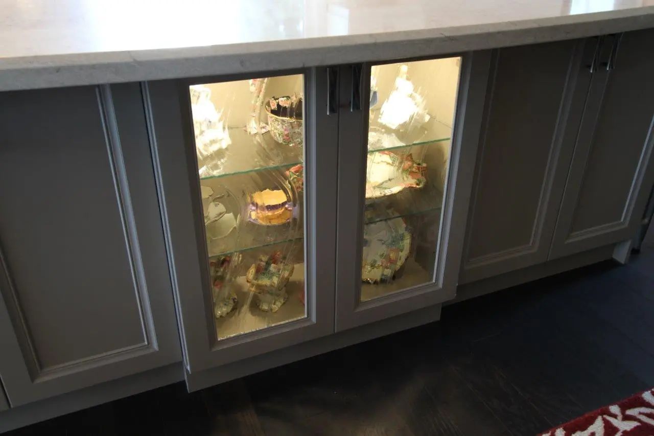Gray kitchen cabinets with a marble countertop, featuring two middle glass-paneled doors displaying illuminated ceramic dishware and decorations inside.
