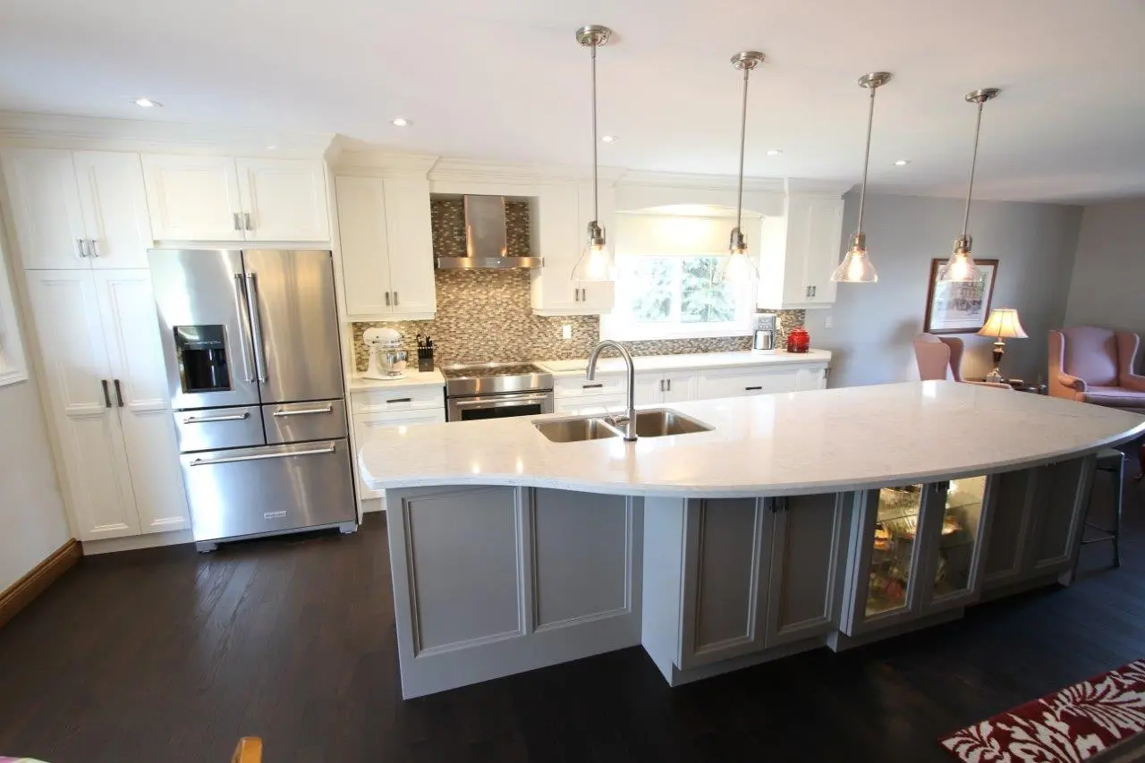 Modern kitchen with a large white marble island countertop featuring a dual sink and hanging pendant lights, white cabinets, stainless steel refrigerator and oven, mosaic tile backsplash, and dark wooden floor.
