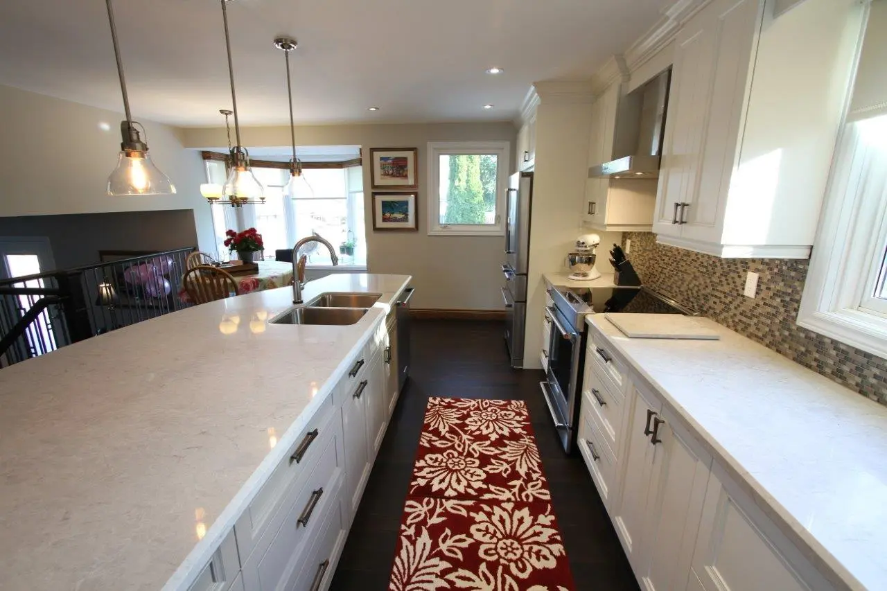 Modern kitchen with white cabinets, marble countertops, stainless steel appliances, a red and white floral runner rug, and pendant lights above an island with a double sink