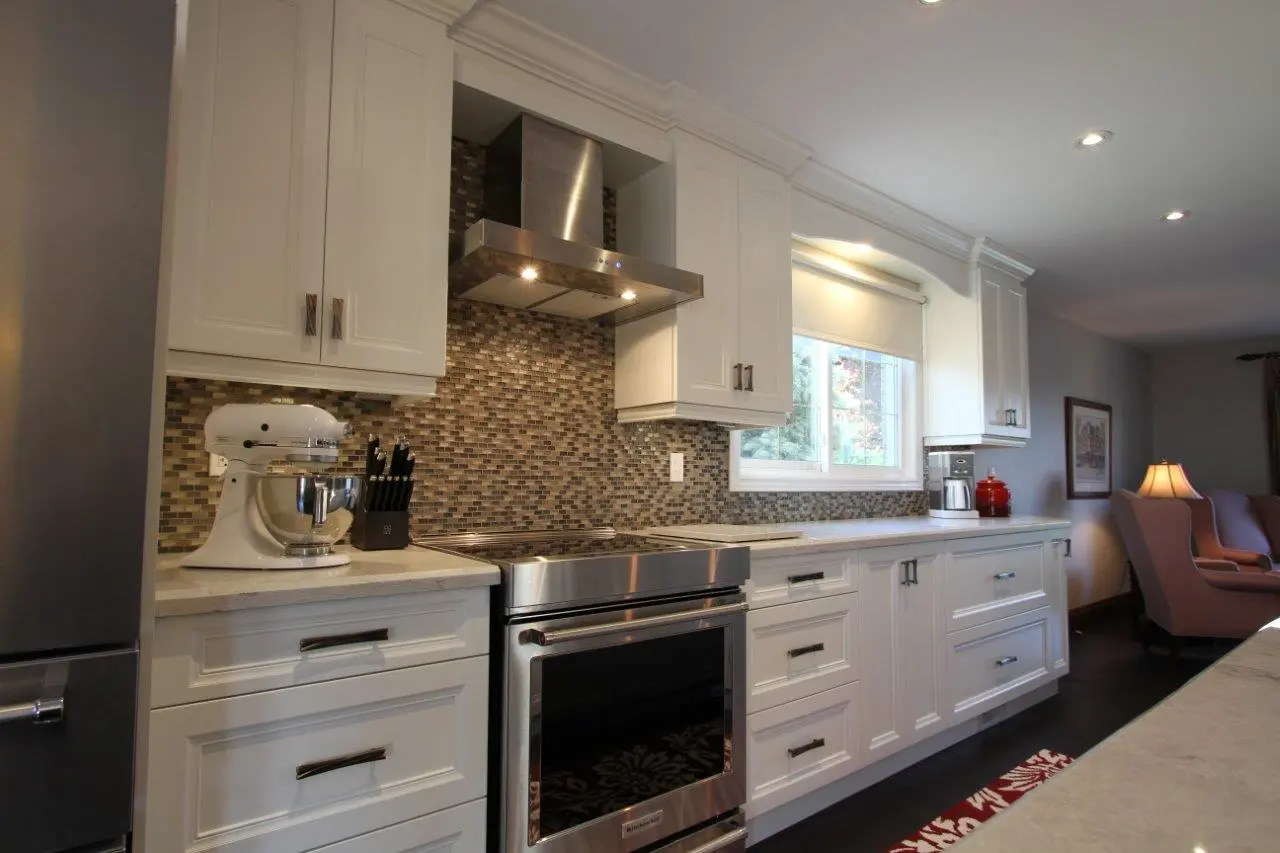 Modern kitchen interior featuring white cabinets, a stainless steel stove with a matching range hood, a mosaic tile backsplash, a window with a white roller blind, a white stand mixer, a knife block, a coffee maker, and a red ceramic container on the counter with soft lighting and a cozy seating area in the background.