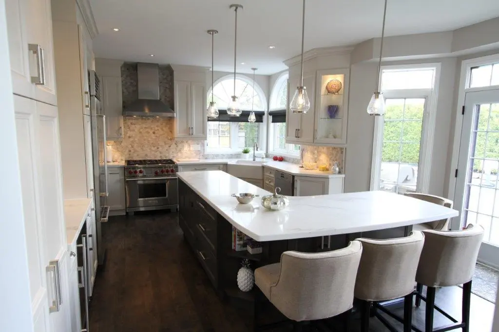 Modern kitchen with a large white marble island, dark wooden base, padded beige chairs, stainless steel stove and range hood, light grey cabinets, and large windows providing natural light.