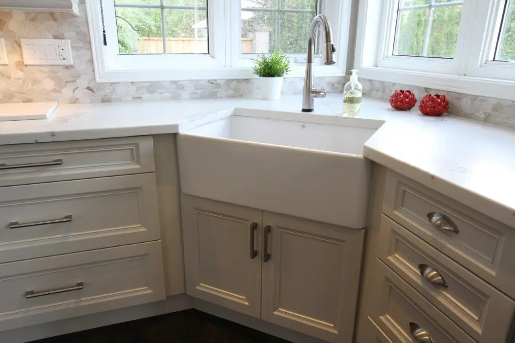 White farmhouse kitchen sink with silver faucet installed under a window, surrounded by white cabinets and countertops, with a small potted plant and soap dispenser on the counter.