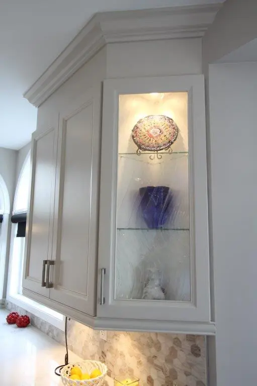 White kitchen cabinetry with a glass display cabinet containing a decorative plate, a blue vase, and a small figurine, illuminated by an overhead light and set above a counter with a mosaic tile backsplash and a bowl of lemons.