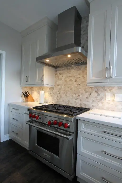 Modern kitchen featuring a stainless steel gas stove with red knobs, a matching stainless steel range hood with lights on, white cabinetry, a knife block, paper towel holder, and a light-colored stone backsplash.