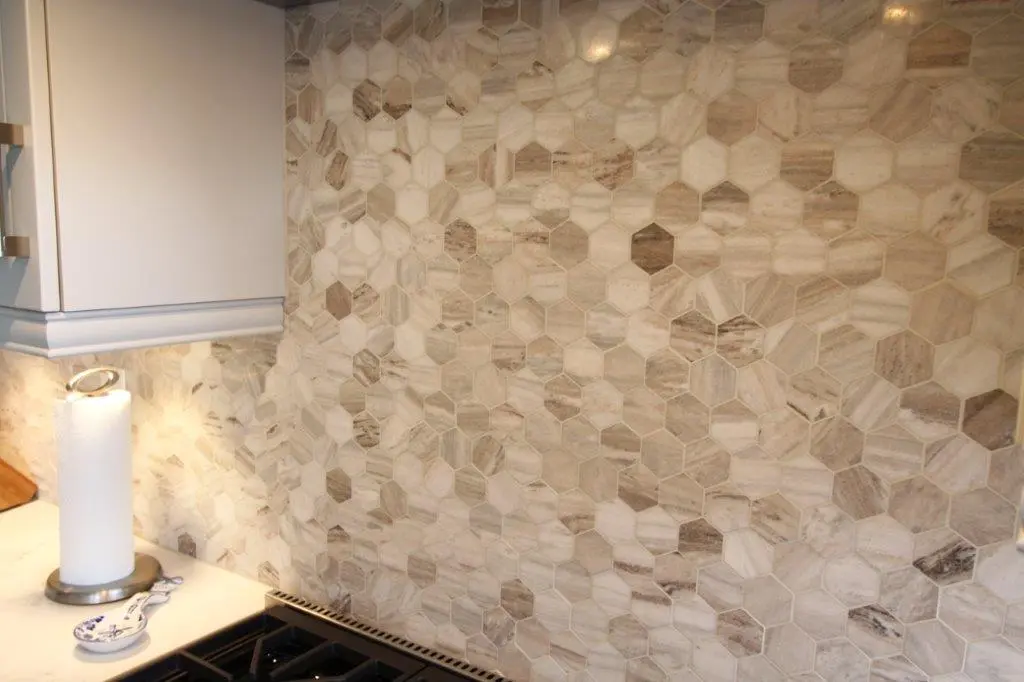 Kitchen backsplash with hexagonal beige and brown tiles, adjacent to a white countertop holding a paper towel holder and a decorative spoon rest, under a white cabinet.